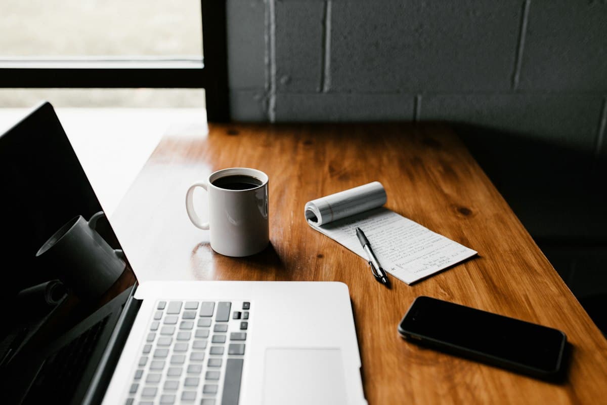 A wooden desk with a laptop, notebook, and coffee in warm light